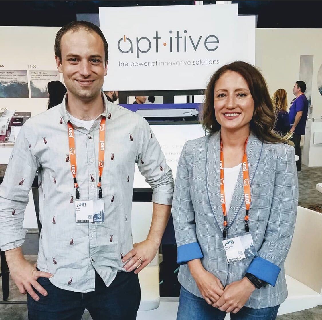 a man and a woman stand in front of an Aptitive booth at a convention